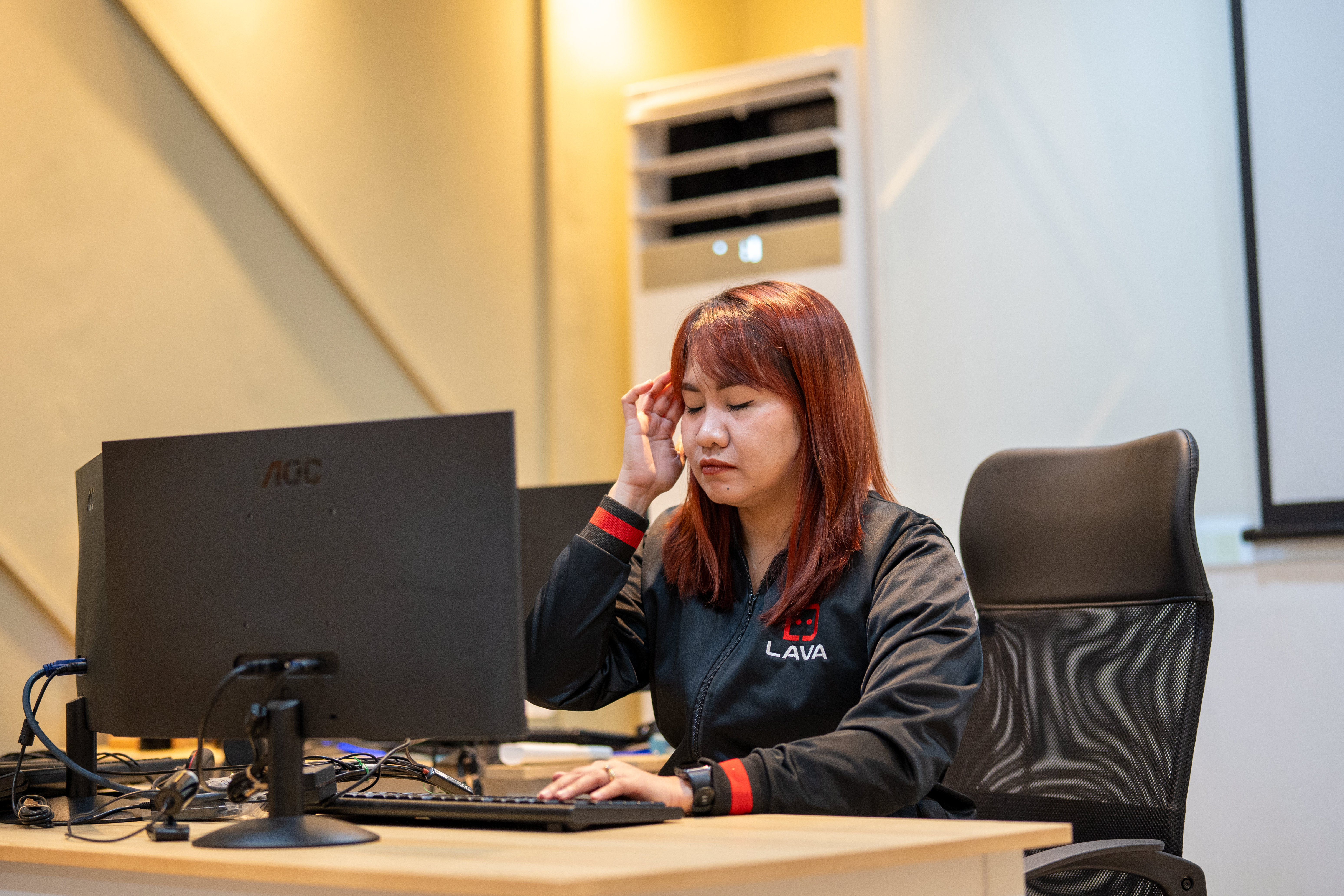 A woman with reddish-brown hair wearing a black and red LAVA branded jacket sits at a wooden desk in an office. She has her eyes closed and one hand raised to her temple