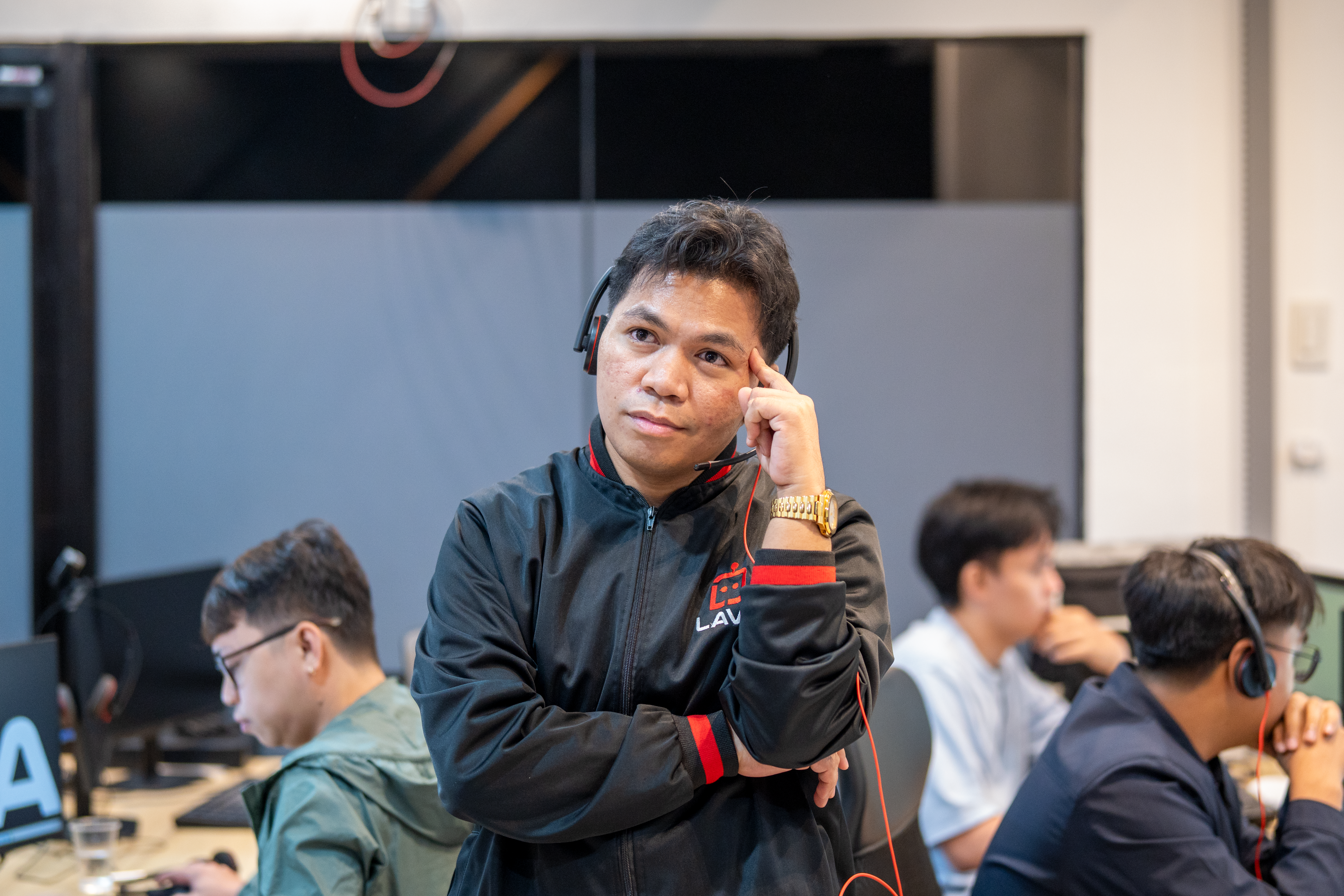 A man wearing a black and red jacket and a headset stands with arms crossed in a gaming or esports facility, resting one hand thoughtfully against his temple. Other participants wearing headsets are seated at computers in the background.