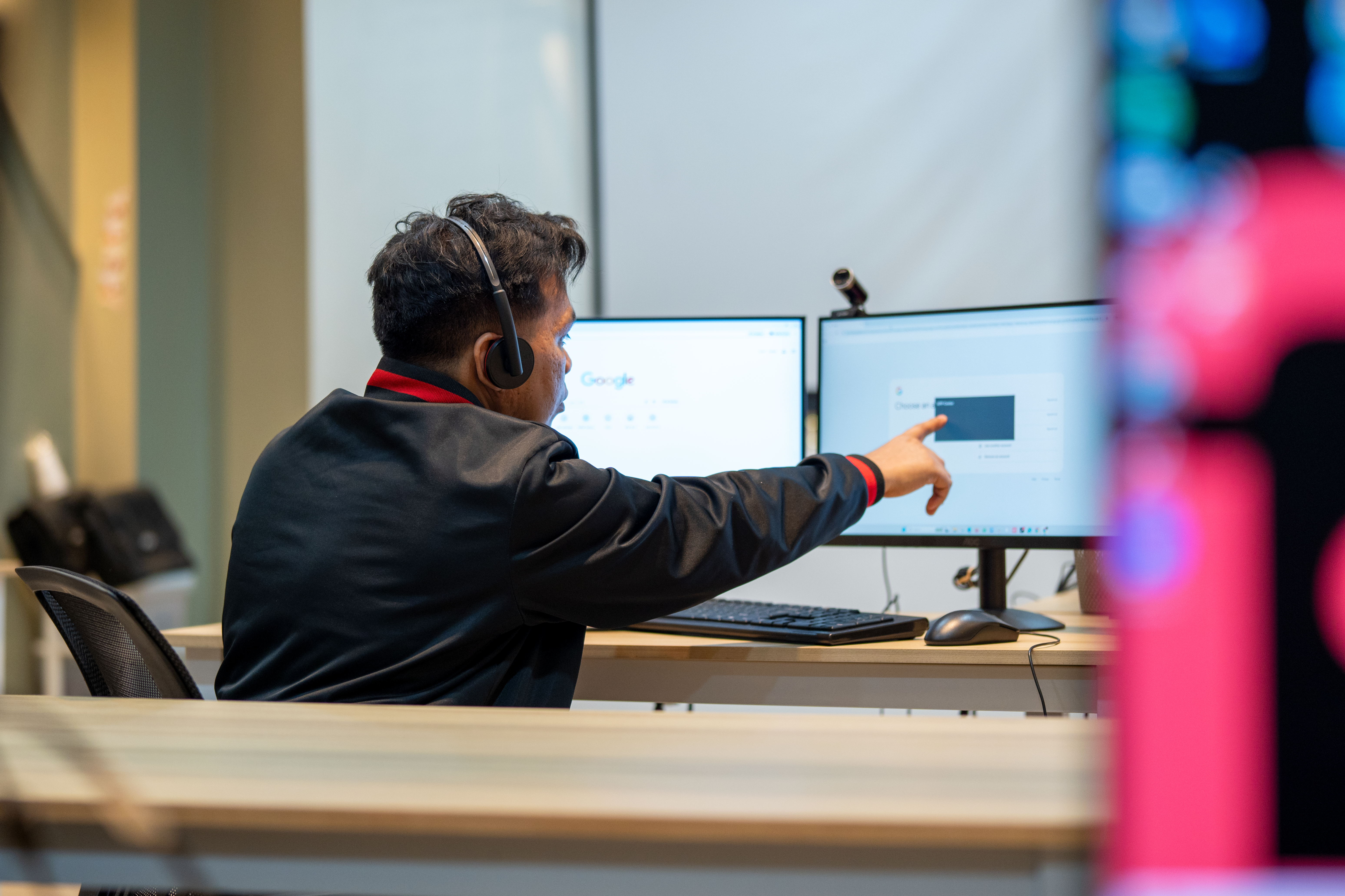 A person wearing a black jacket and headset sits at a desk in an office or esports facility, pointing at one of two monitors displaying a Google Chrome browser. A keyboard, mouse, and webcam are visible on the desk.