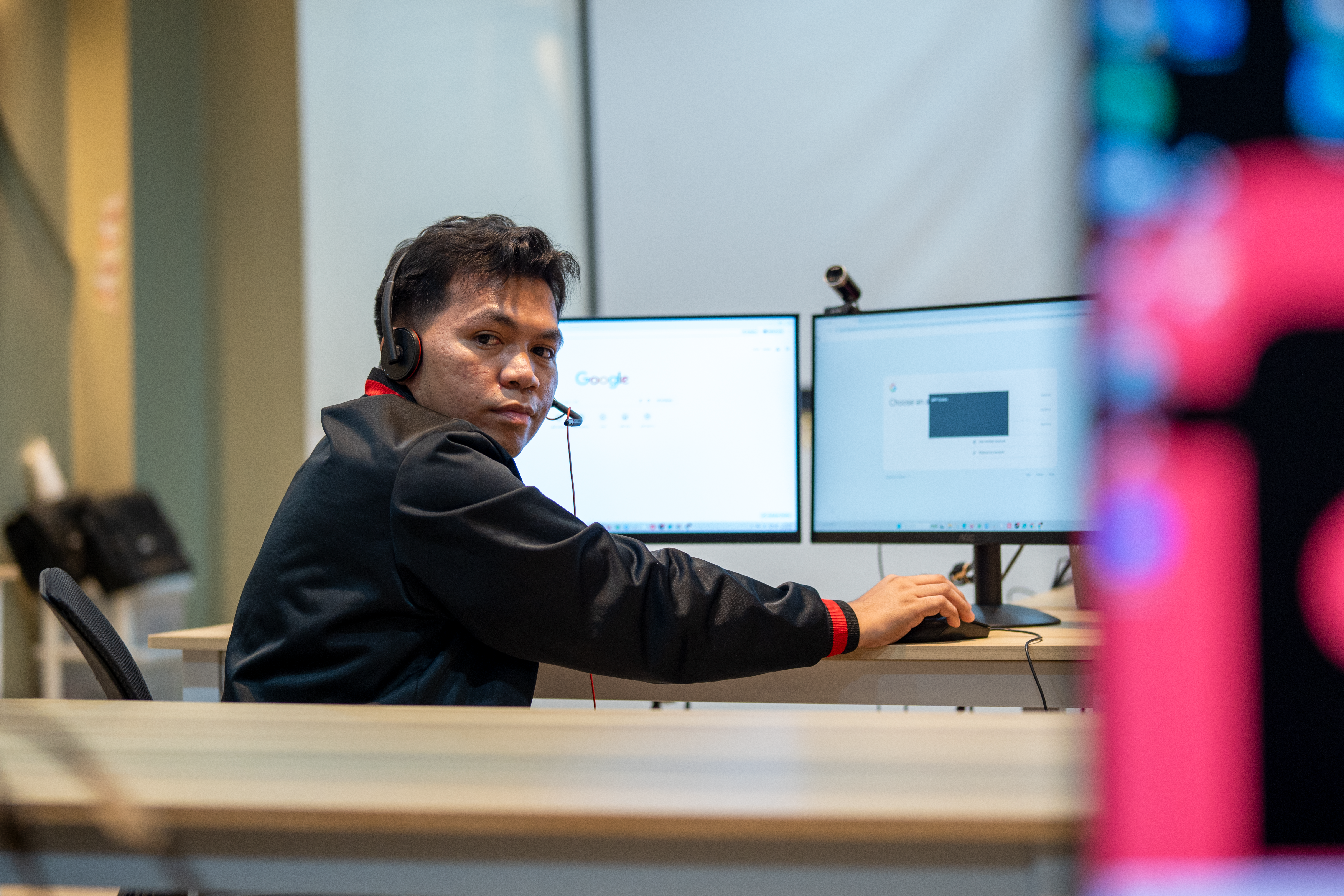 A man wearing a headset looks over his shoulder while working at a dual-monitor computer setup in an office.
