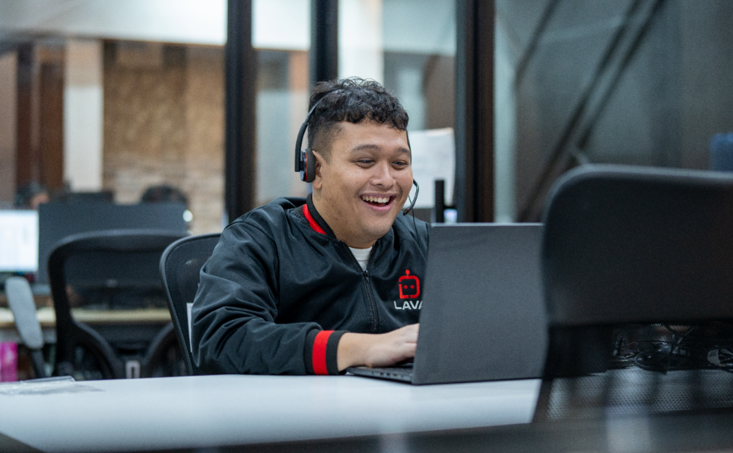 A man wearing a headset and a black LAVA-branded jacket smiles while typing on a laptop at an office desk, with blurred office workstations in the background.