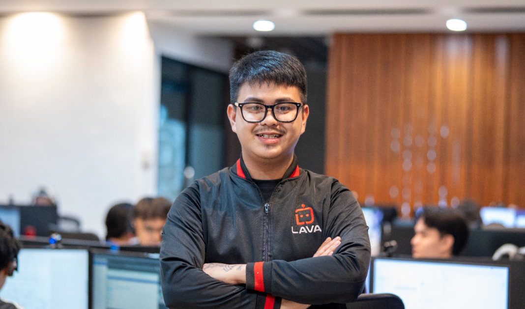  man wearing glasses and a black LAVA-branded jacket stands with arms crossed, smiling at the camera, with colleagues working at computer monitors in the background.