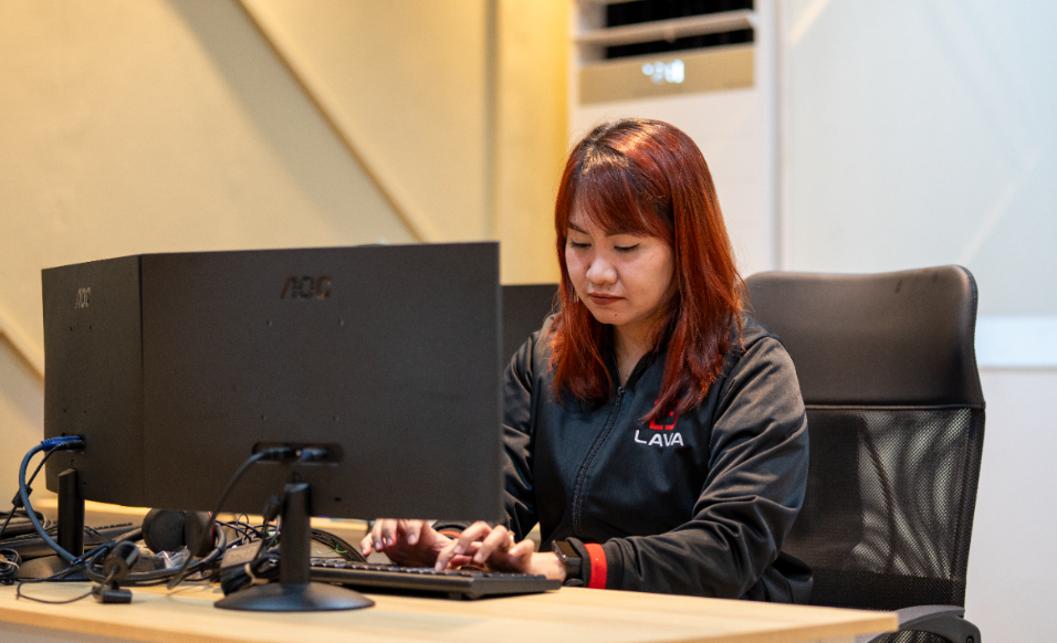 A woman with red hair wearing a black LAVA-branded jacket types on a keyboard while looking at a monitor, seated at a desk with cables and a second monitor visible.