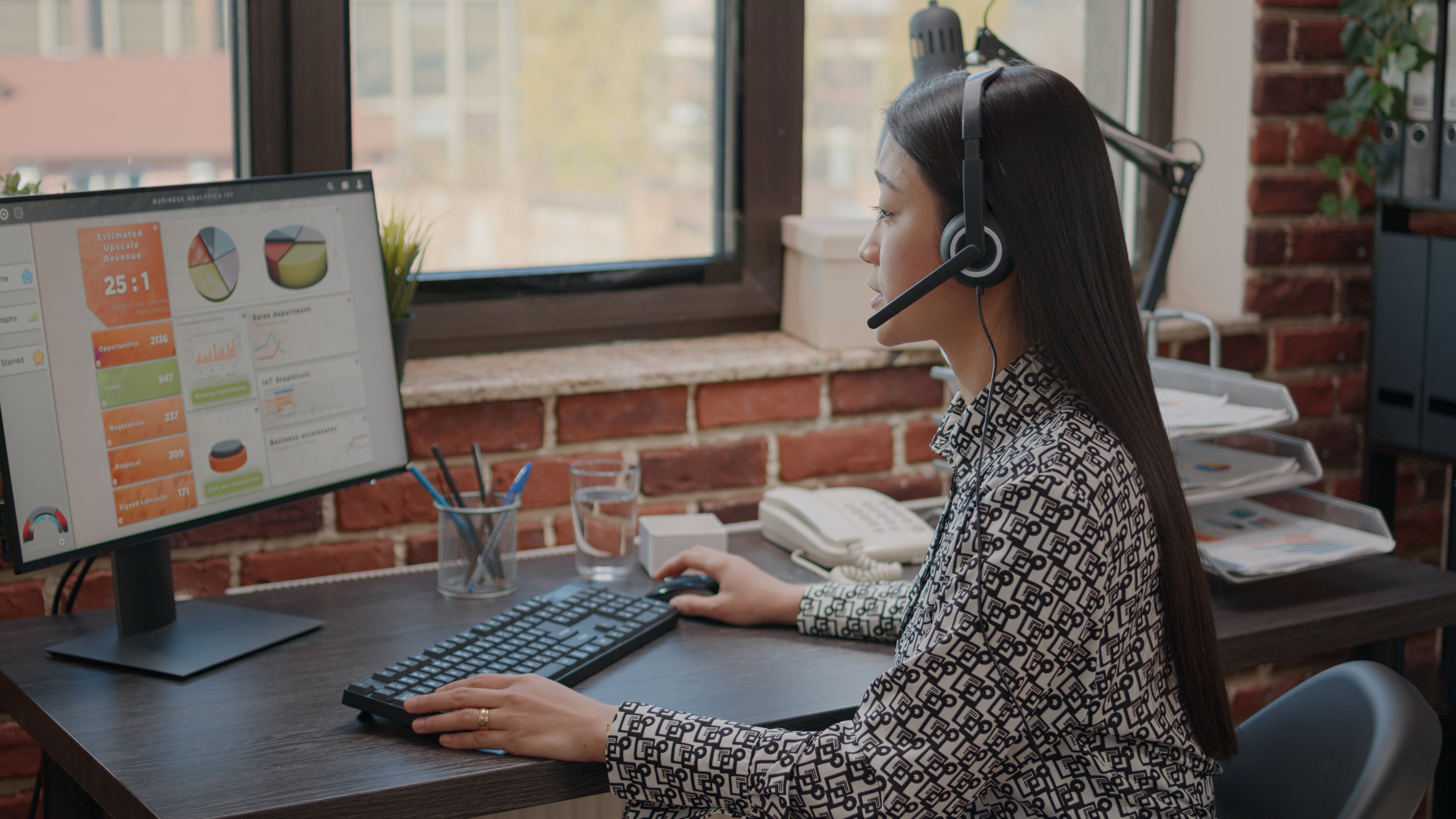 A woman with long dark hair wearing a headset sits at a desk, working at a desktop computer displaying a business analytics dashboard with charts and metrics. She is in a modern office with brick walls and large windows.