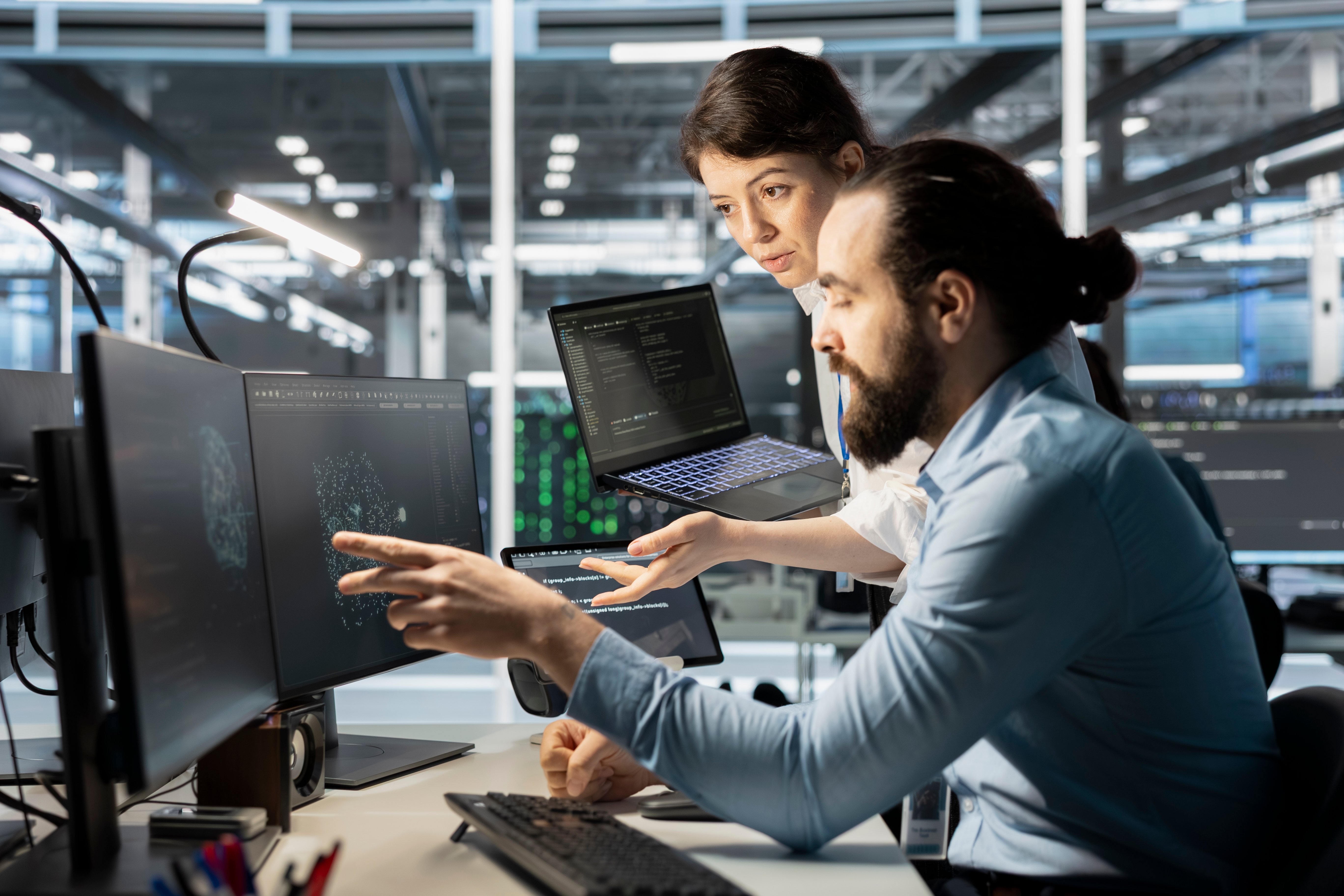 Two tech professionals collaborate at a multi-monitor workstation in a modern data center, with one pointing at a screen displaying a glowing network visualization while the other looks on attentively.