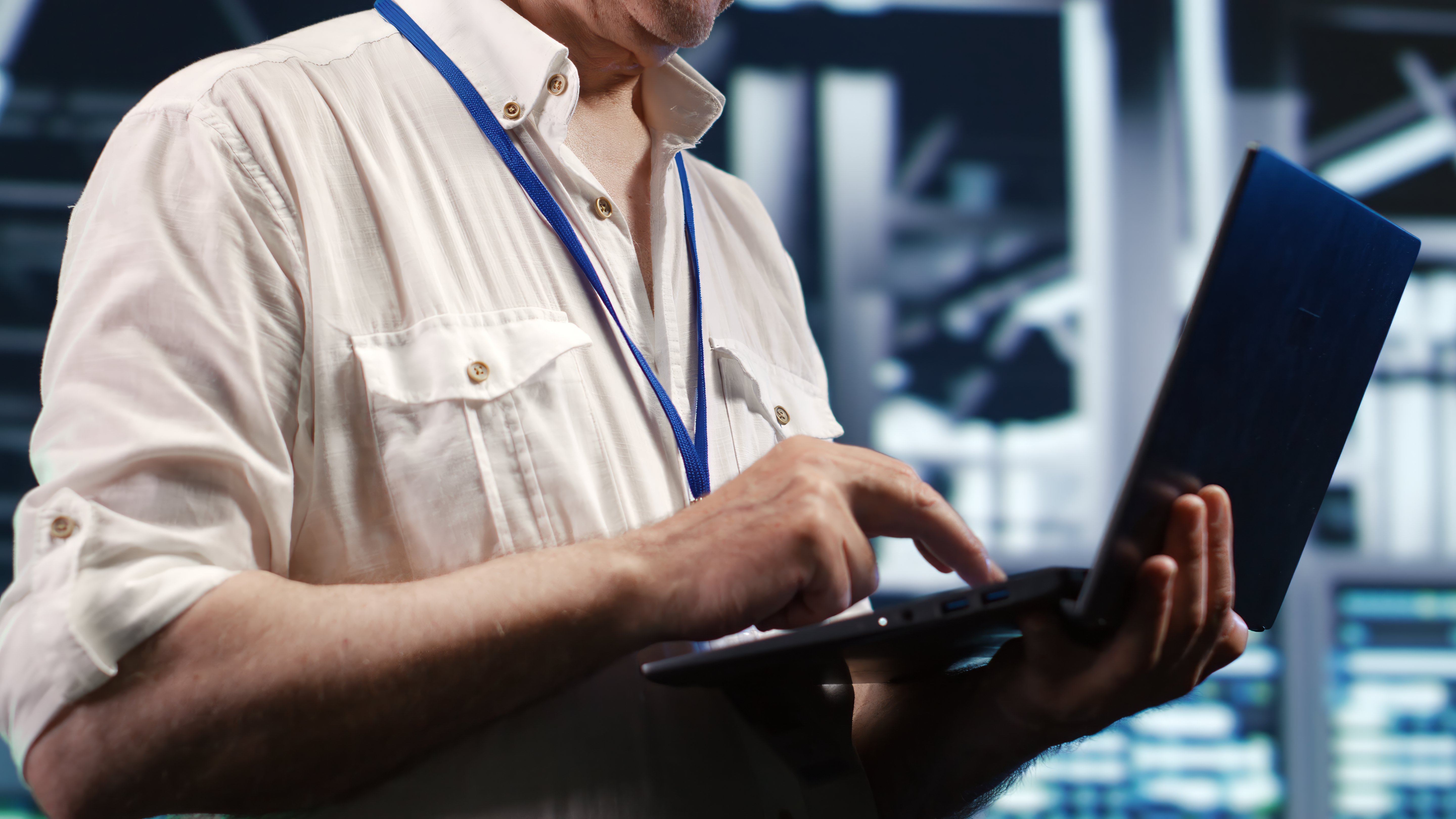 IT professional wearing a blue lanyard stands in a server room, typing on a laptop he holds in his arms.