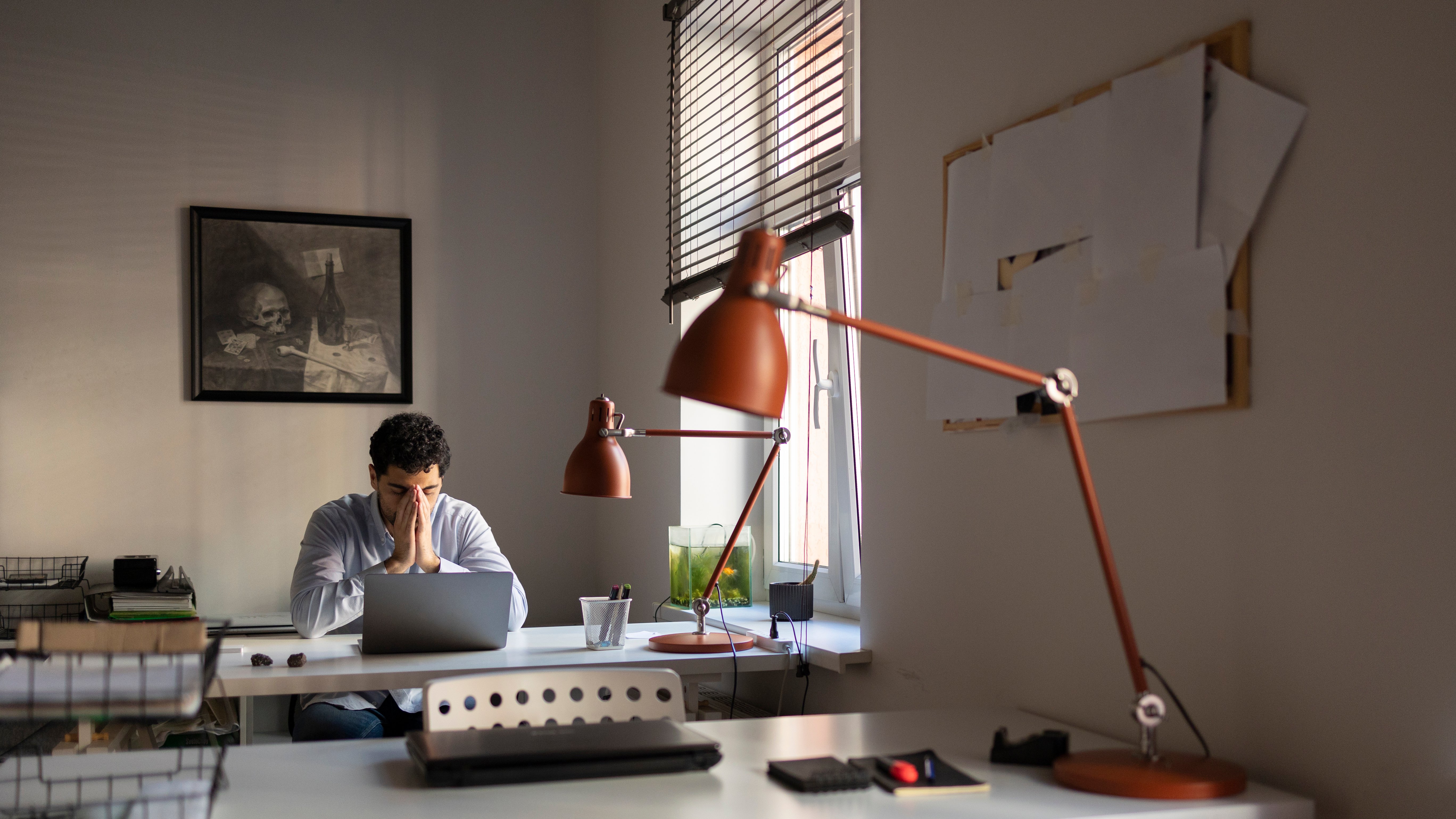 A stressed man sits at a desk with his head in his hands, elbows resting on either side of an open laptop, in a dimly lit office. A large orange articulated desk lamp dominates the foreground, unlit, alongside a small fish tank on the windowsill and blueprint-style papers pinned to the wall. A framed vanitas-style still life painting