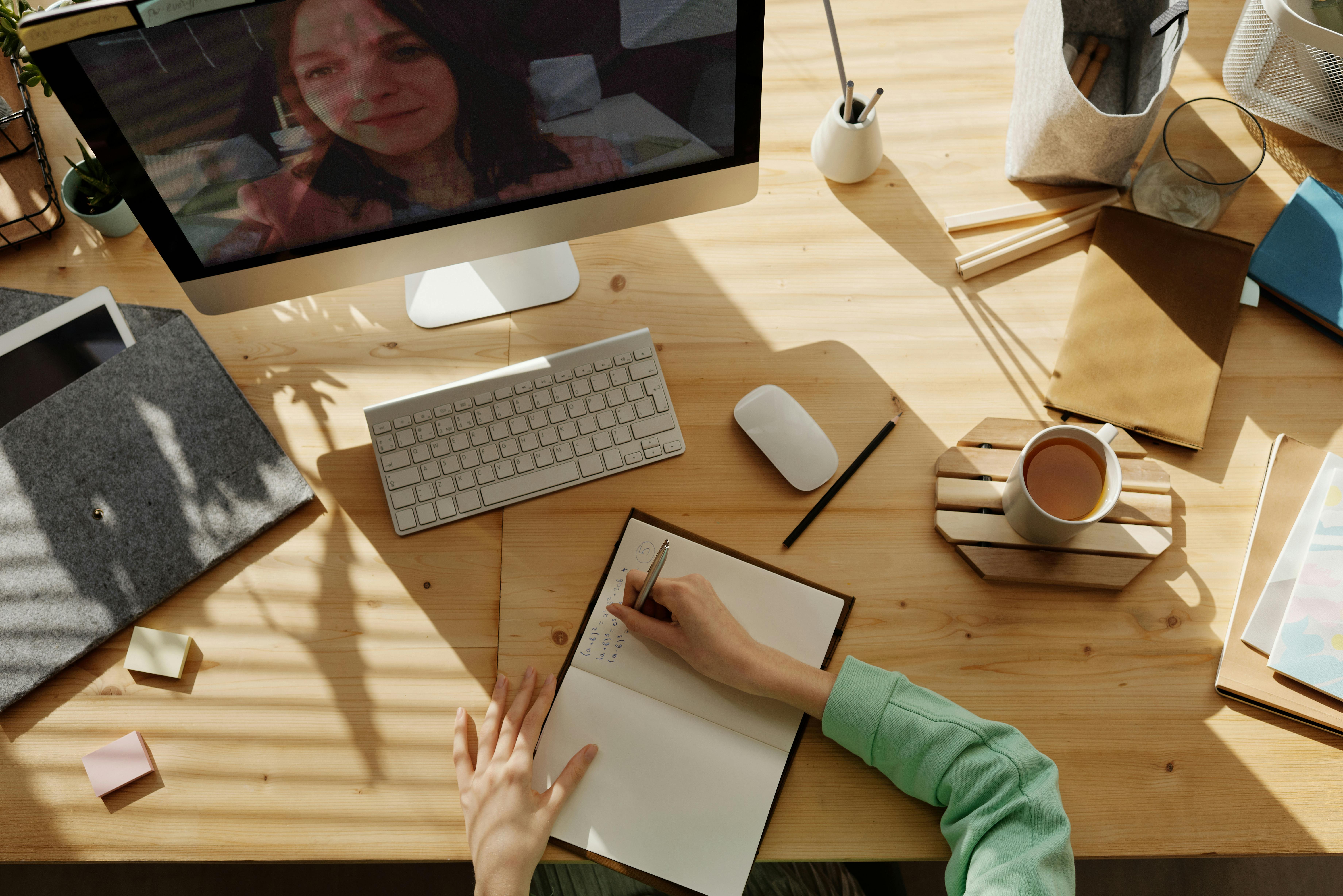 Person on a virtual meeting taking notes at a desk with computer, coffee, and office supplies.