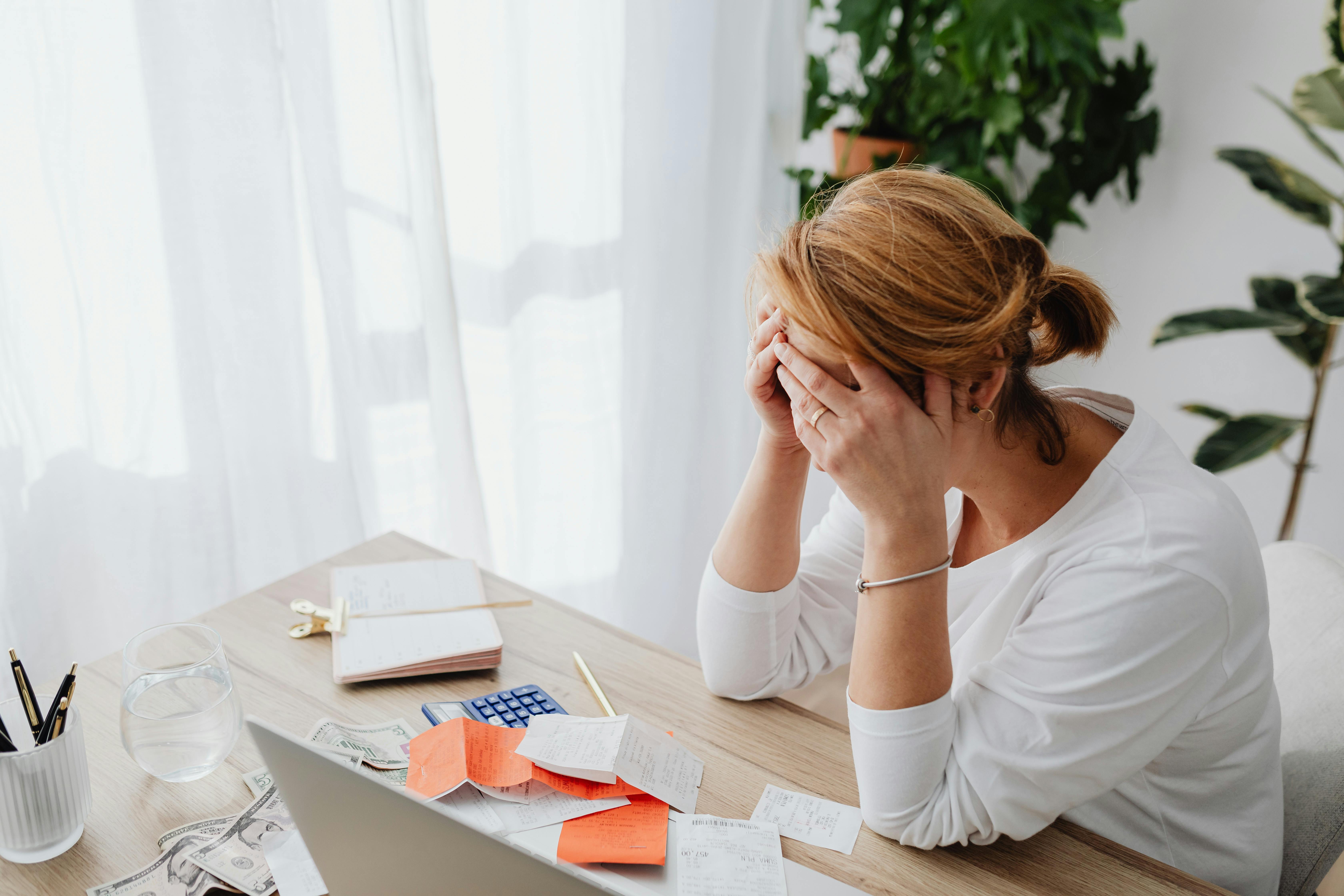 Stressed office worker holding head in hands at desk cluttered with receipts and papers.