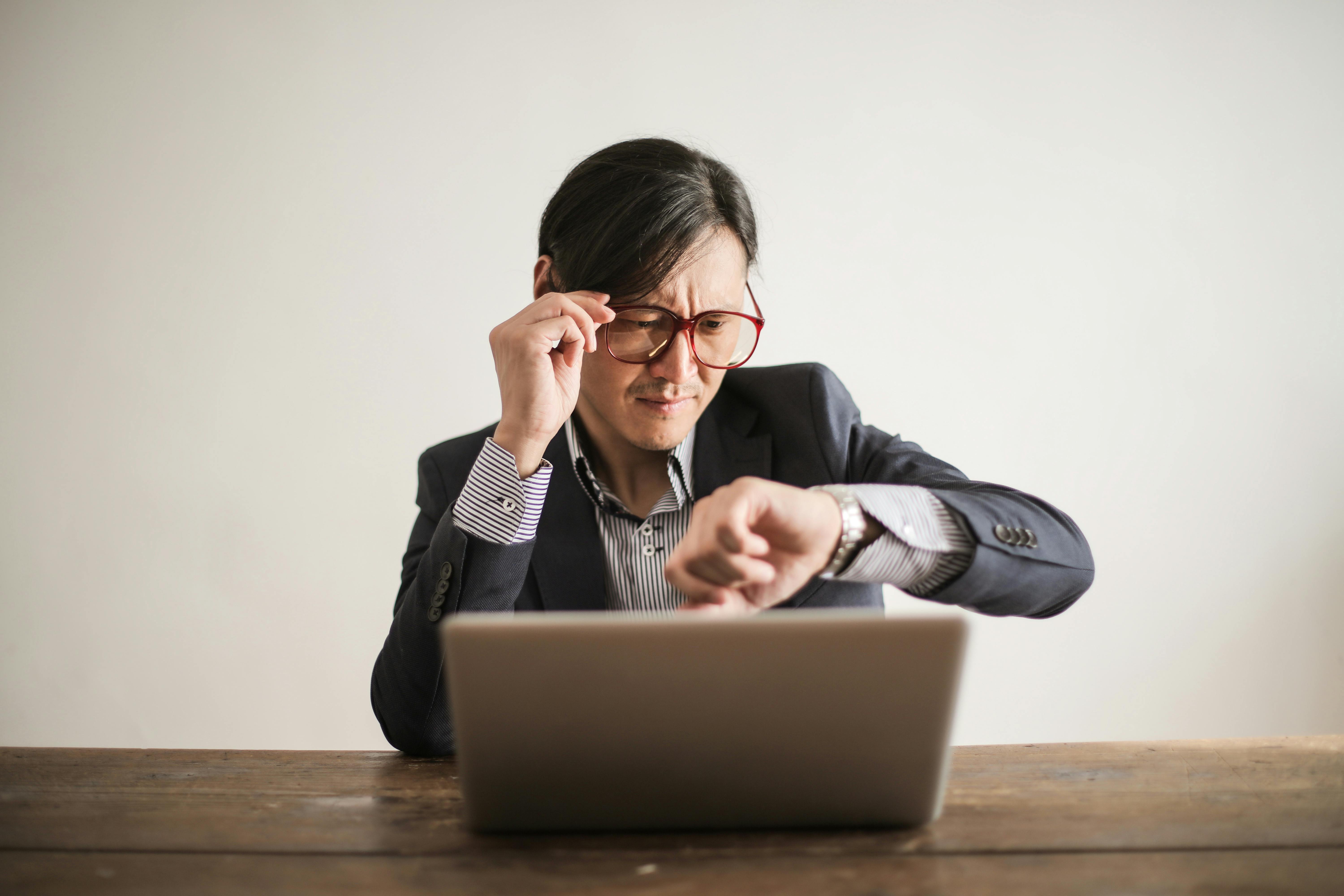 businessman checking wrist watch over a laptop, representing a deadline, urgency, or time management issue.