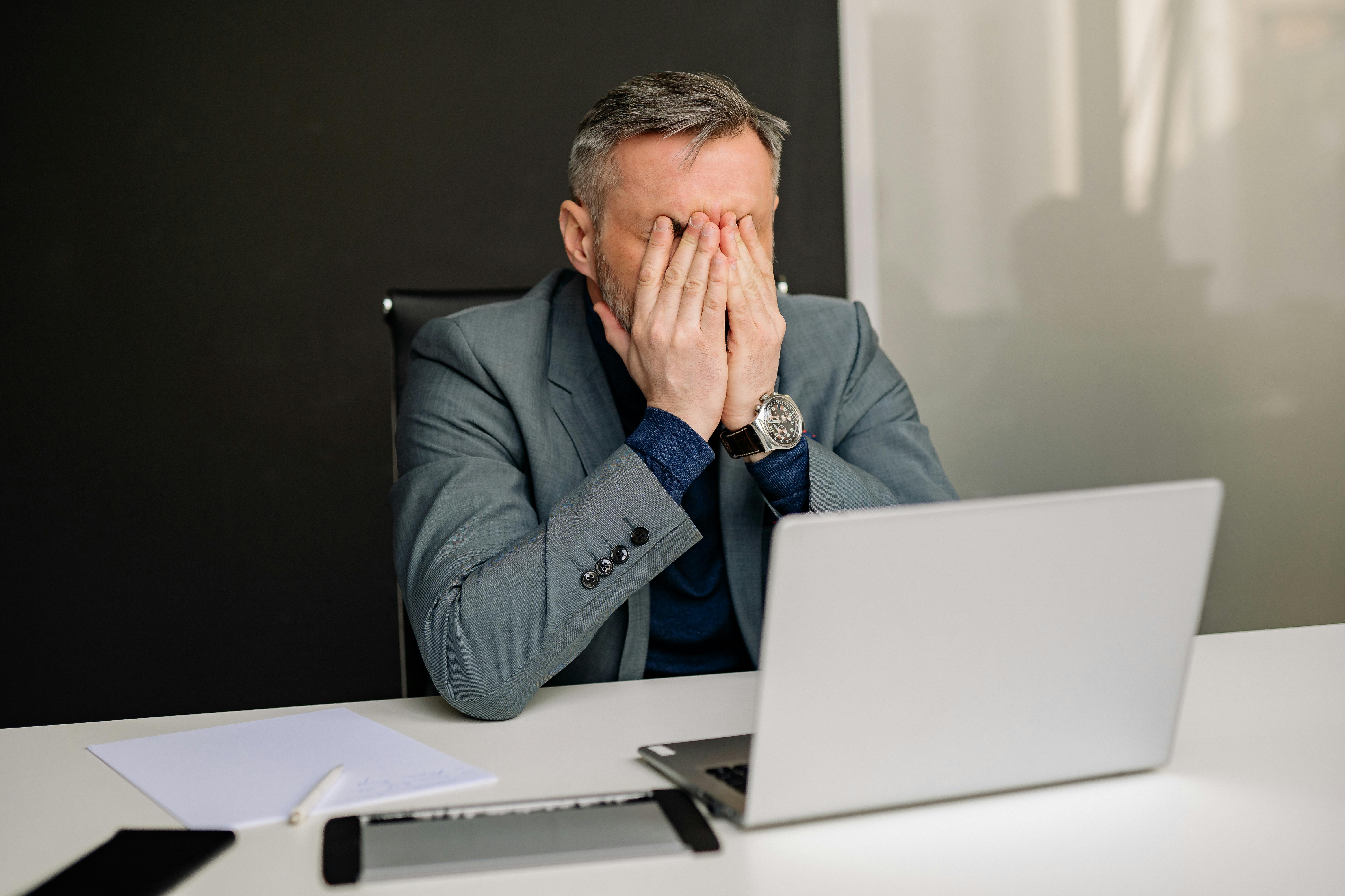 Frustrated businessman in a suit sitting at a desk with a laptop covering his face with his hands.