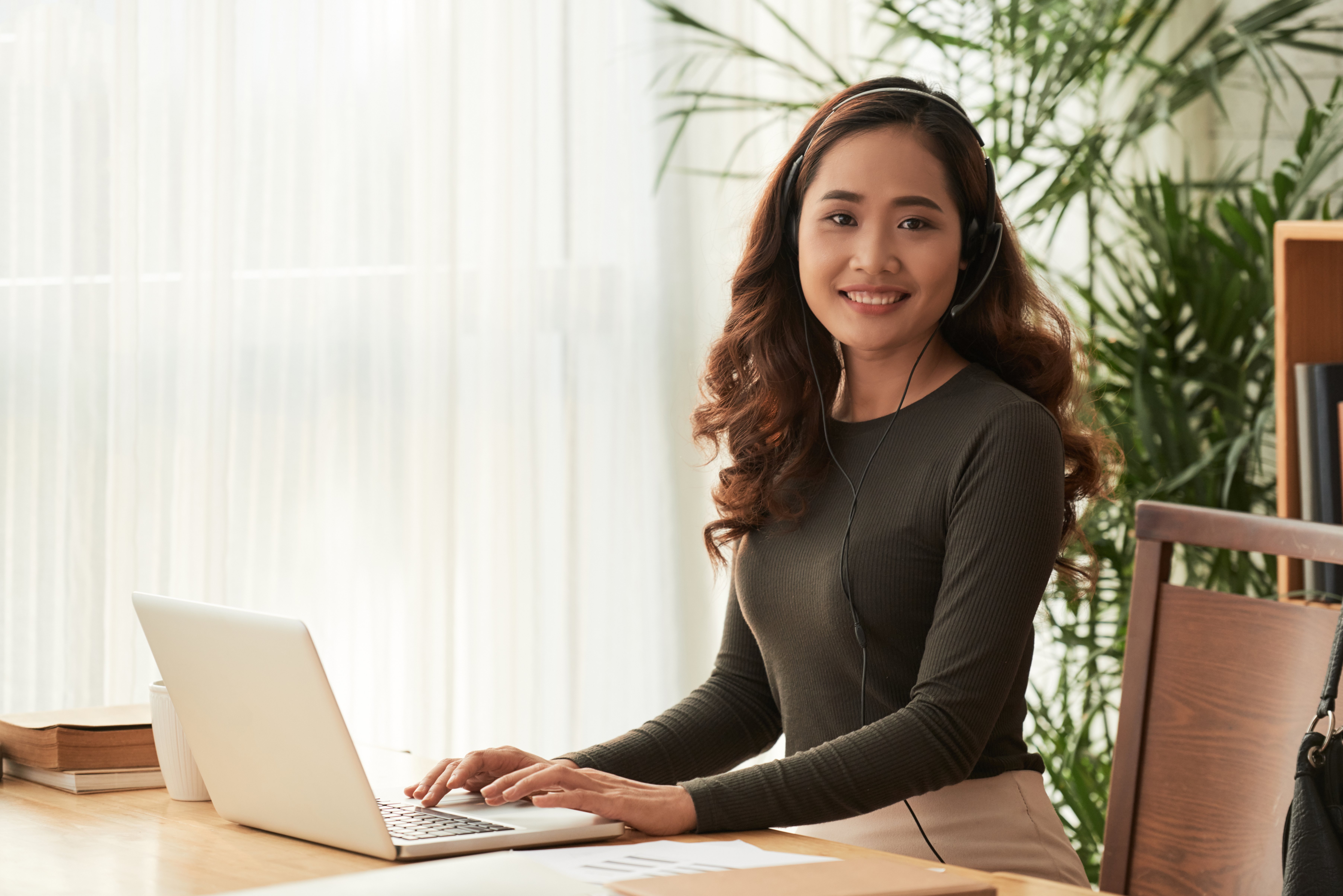 A smiling young woman with curly auburn hair wearing a headset sits at a wooden desk with an open laptop in a bright, plant-filled workspace.