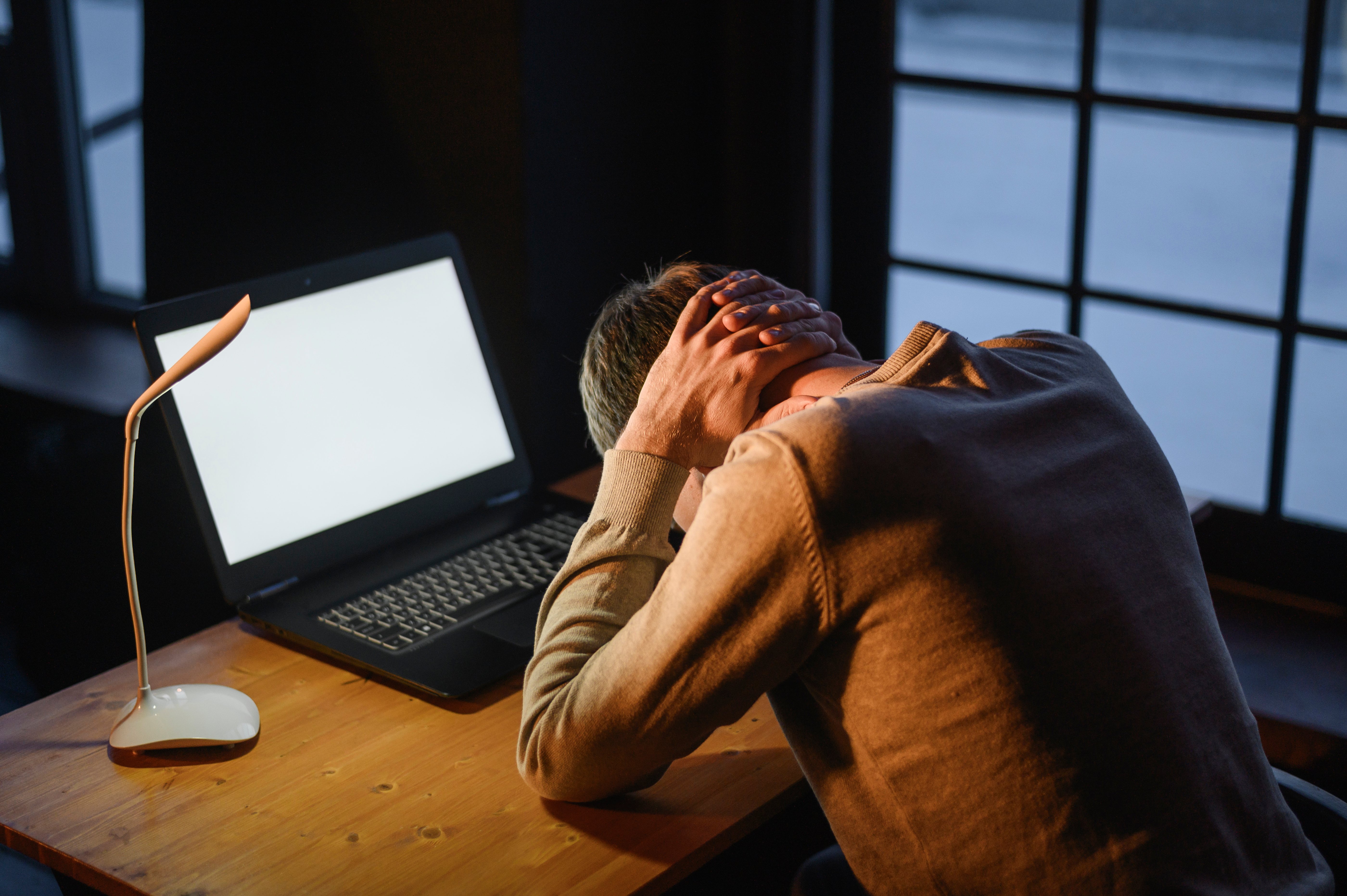 Man hunched over laptop at night, holding his head in stress or exhaustion.