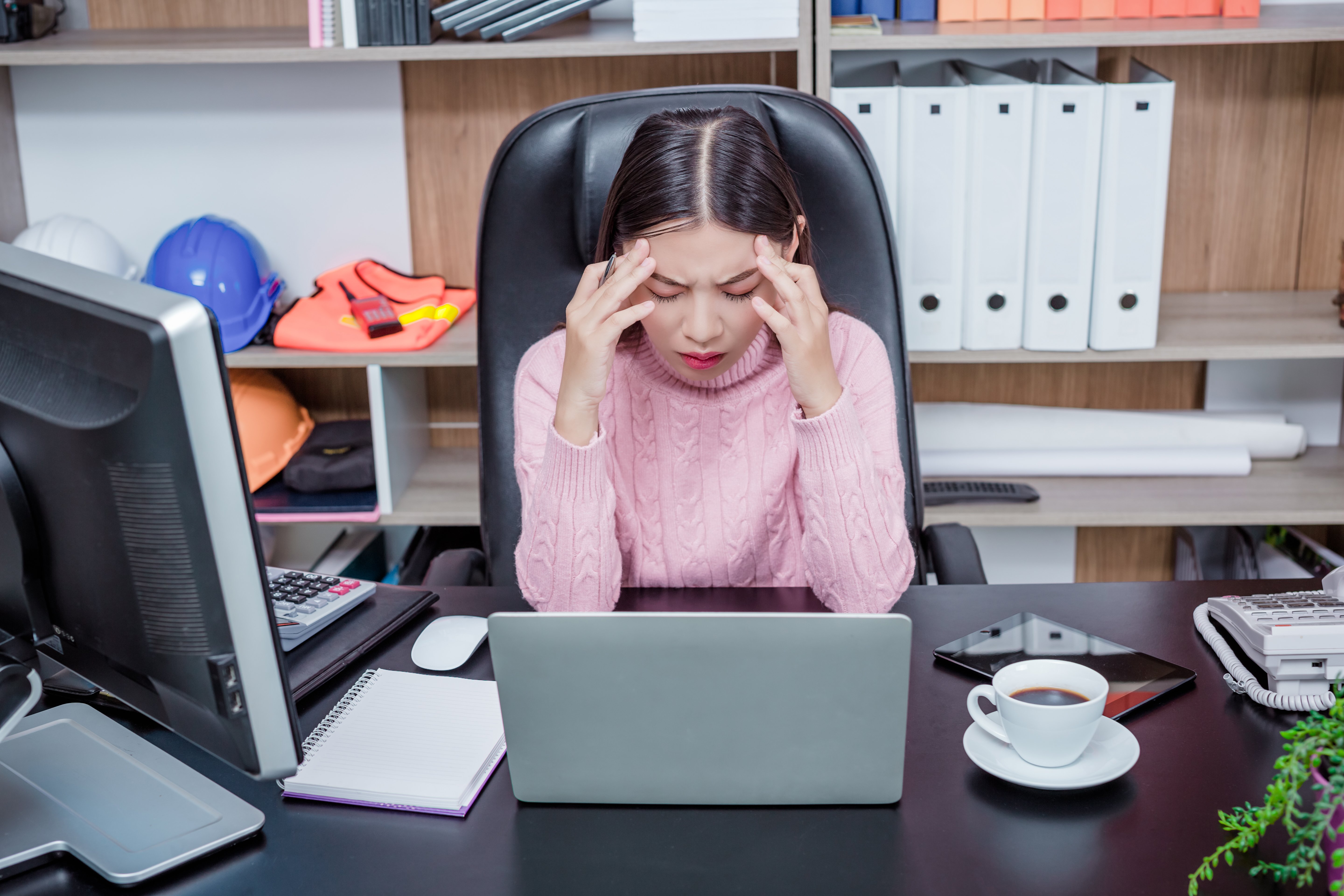  A young woman in a pink sweater sits at her office desk pressing her temples in visible stress, surrounded by a laptop, coffee, and safety equipment on shelves behind her.
